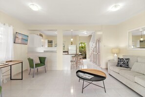 A modern living room with a sofa, a coffee table, a dining area with stools, a kitchen in the background, and a large window. - Palm View 4 - Cozy 2 bedroom apartment in Barbados (Bridgetown)