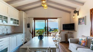 Eine moderne Küche mit weißen Schränken, ein Essbereich mit Tisch und Stühlen, ein Fernseher und Blick aufs Meer.