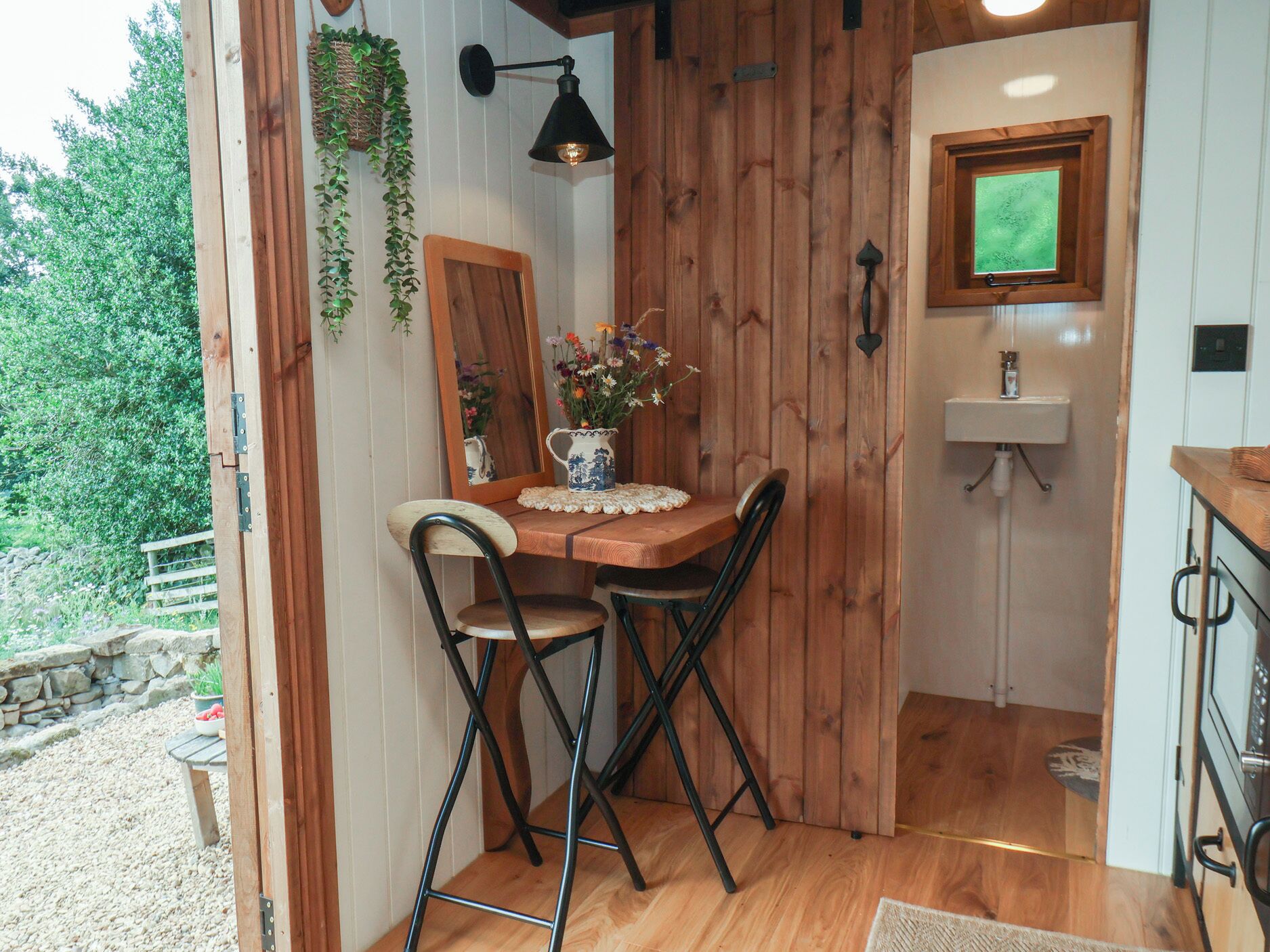 A wooden dining table with two chairs, a vase of flowers, a hanging plant, and a wall-mounted light fixture.