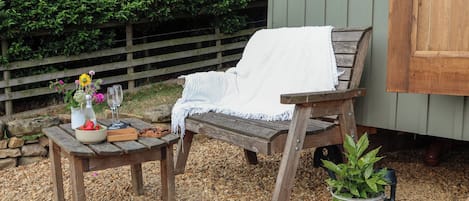 Un área de estar al aire libre con bancos de madera, una mesa con un bol de fruta, una botella y vasos.