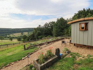 Ein rustikales Holzhäuschen mit Stein Fundament, einer Holzterrasse und Blick auf sanfte Hügel sowie eine eingezäunte Weide.