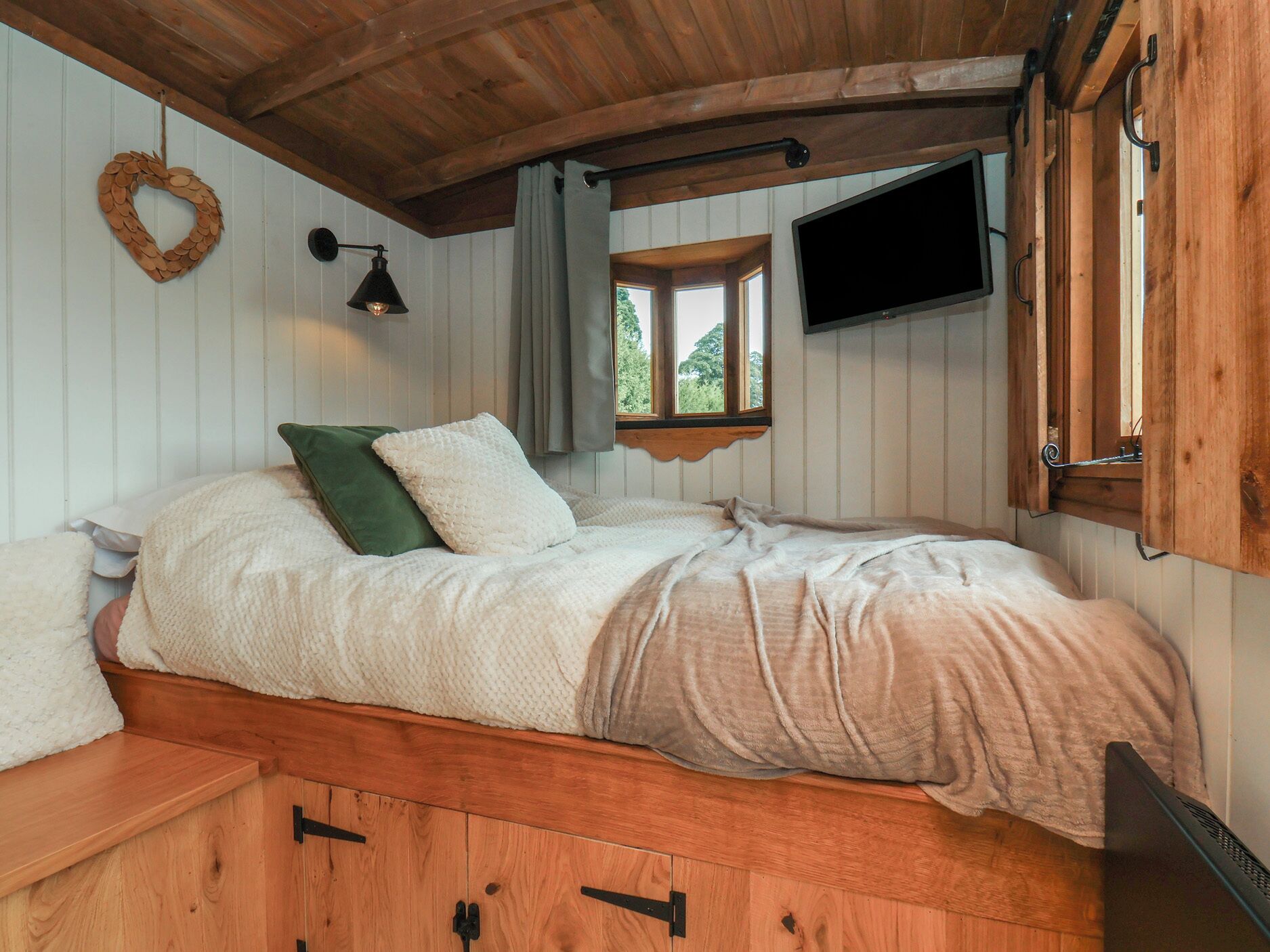 A wooden bed with a headboard, a wall-mounted TV, and a window with curtains.
