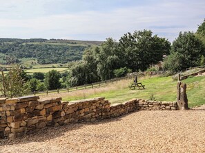 Un muro de piedra, un sendero de grava y una mesa de picnic en un entorno rural con vistas a colinas y árboles.