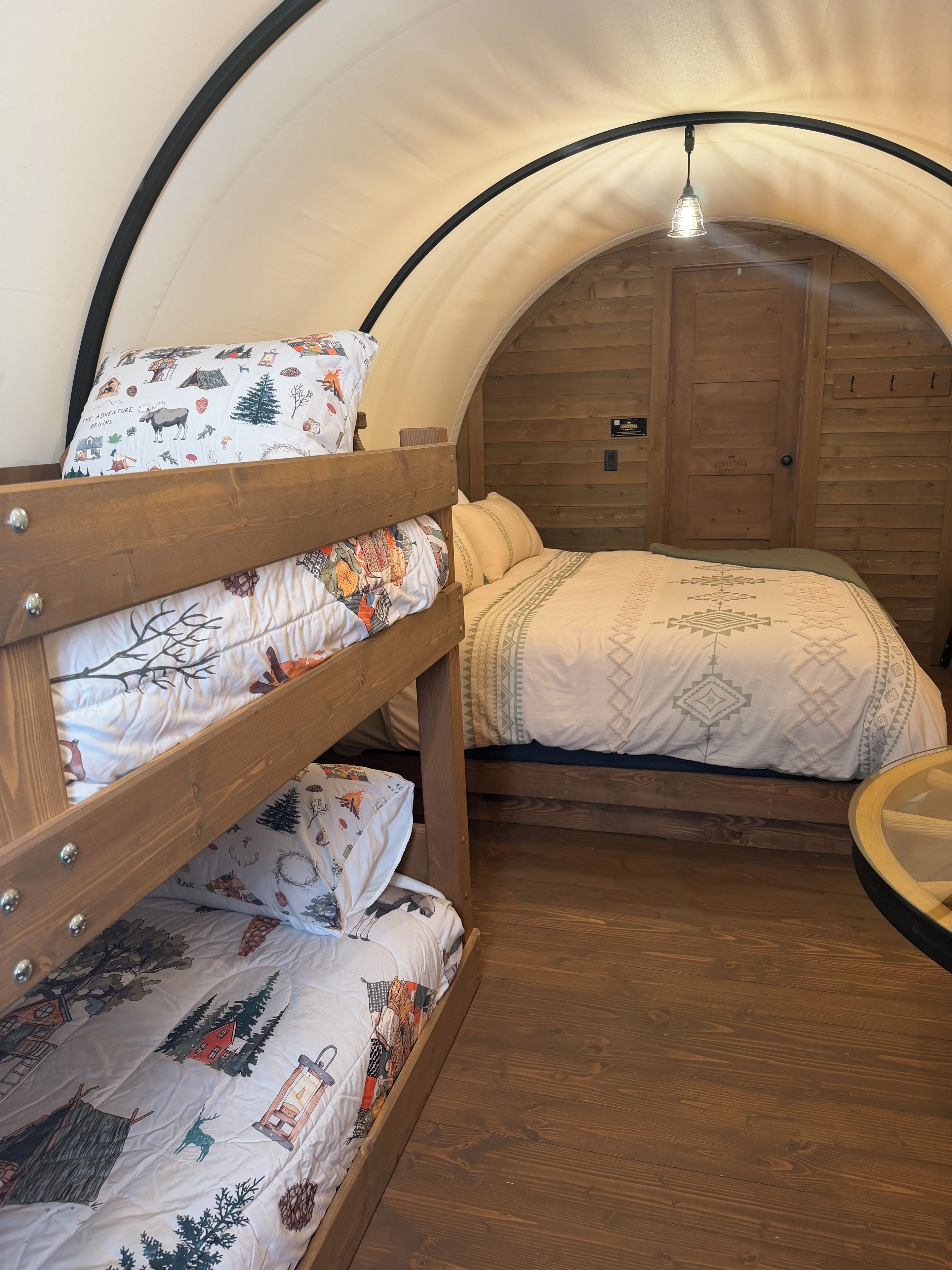 Un lit superposé avec une literie à motifs et un lit simple avec une couvre-lit à motifs, dans une pièce ressemblant à une tente, avec des murs en bois et une porte portant l’inscription « CONESTOCK ».