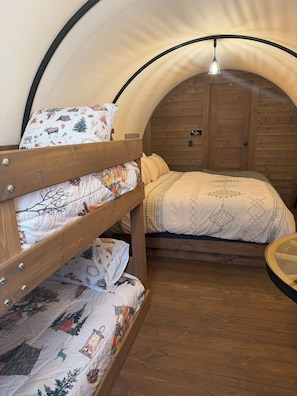 Un lit superposé avec une literie à motifs et un lit simple avec une couvre-lit à motifs, dans une pièce ressemblant à une tente, avec des murs en bois et une porte portant l’inscription « CONESTOCK ».