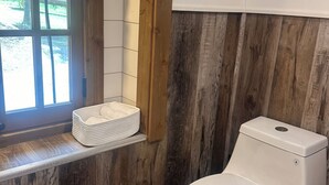Ett badrum med en väggpanel av trä, en vit toalett och ett fönster med en korg på fönsterbrädan.