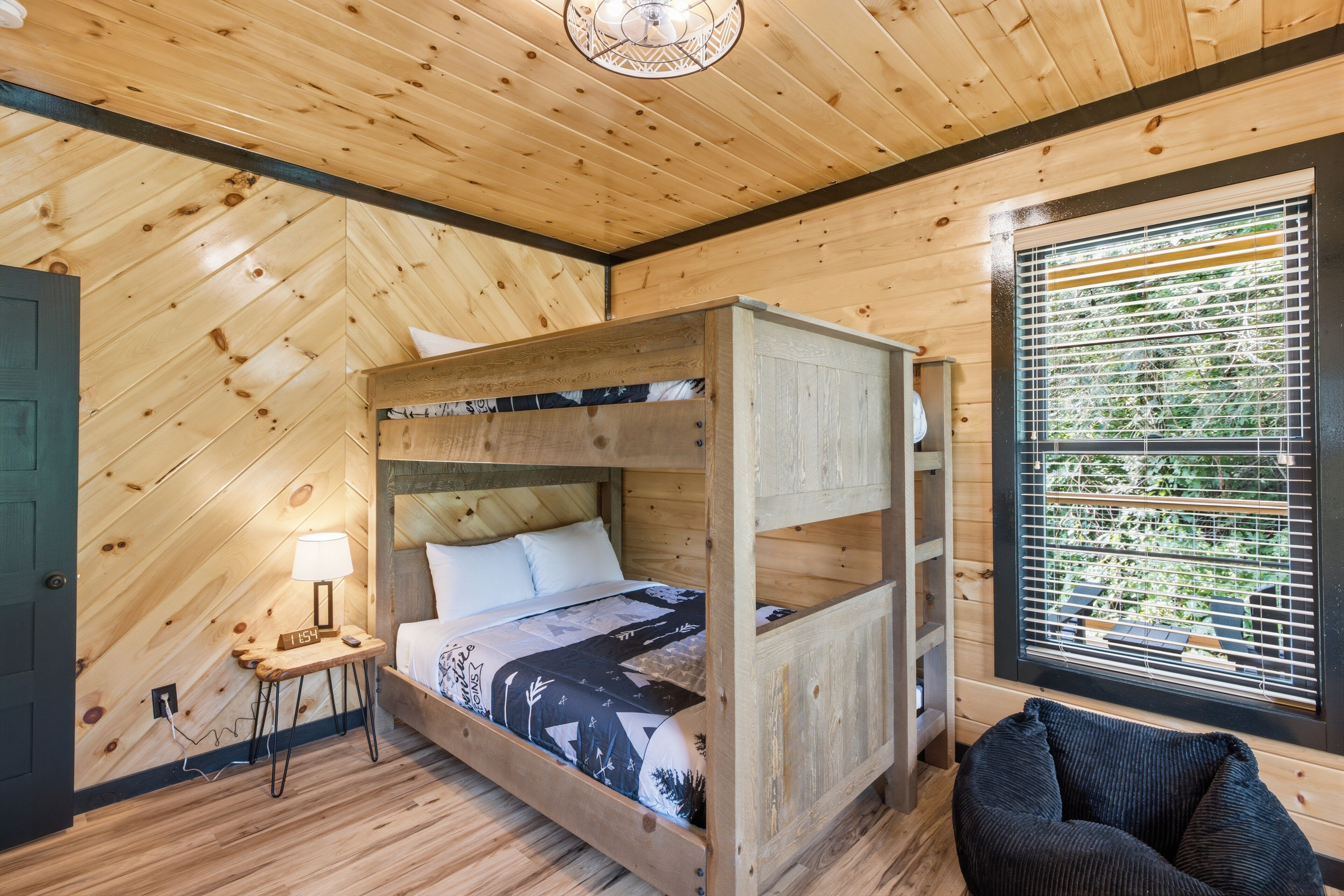 Una habitación tipo cabaña de madera con una litera, una mesita de noche con lámpara, una silla y una ventana con persianas.