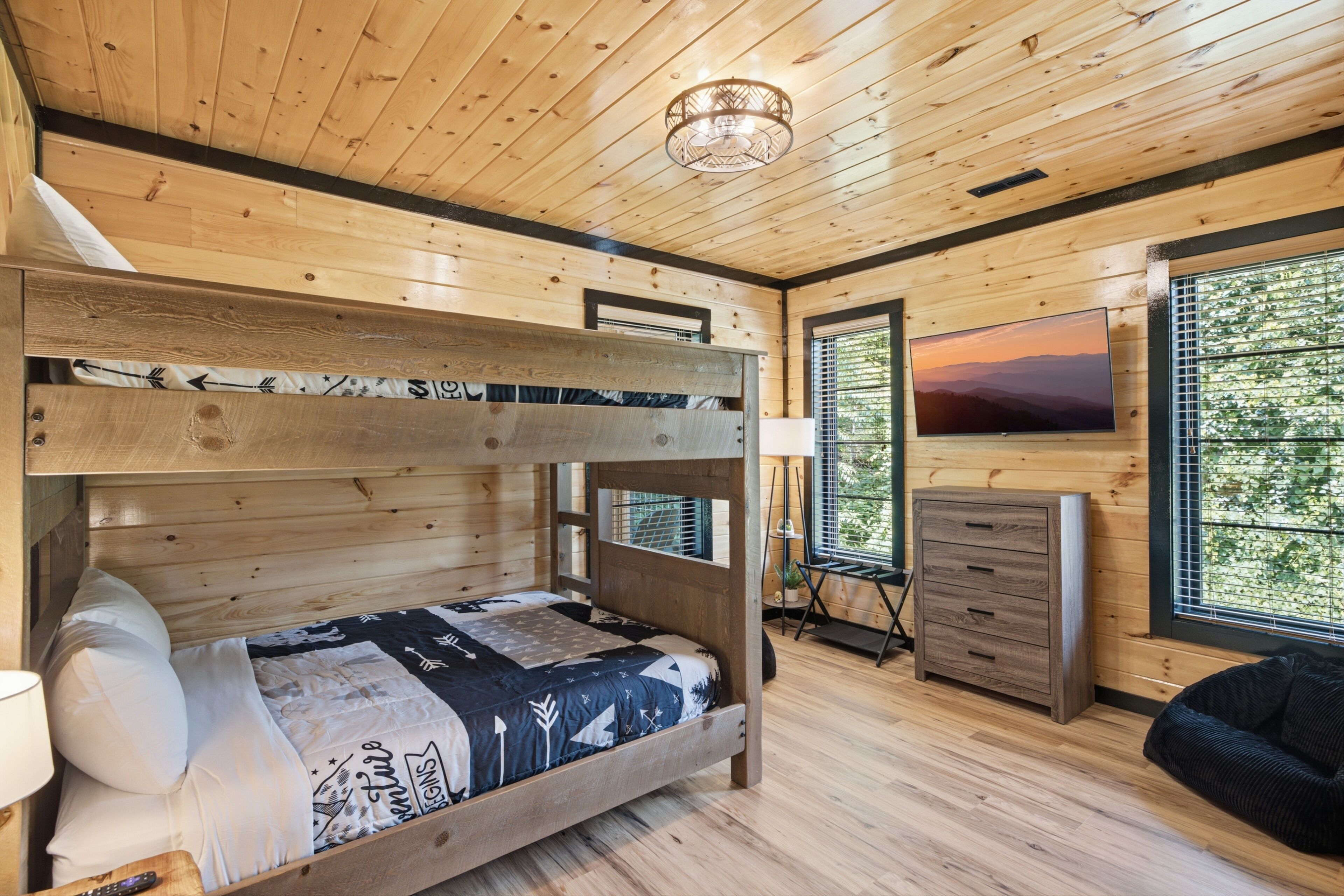 A wooden cabin interior with a bunk bed, a TV, a dresser, and a window with a view of trees.