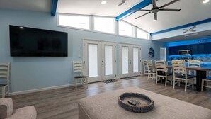 A modern living room with a flat-screen TV, a dining area with chairs, a kitchen with blue cabinets, and a ceiling fan. - New 5br Remodel, Heated Pool, Gameroom, Up To 20 4 Bedroom Home (South Padre Island)