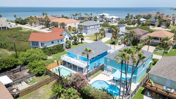 A residential area with a large blue house featuring a swimming pool, surrounded by palm trees and a view of the ocean. - New 5br Remodel, Heated Pool, Gameroom, Up To 20 4 Bedroom Home (South Padre Island)