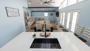A modern kitchen with a central island, a dining area with a long table and chairs, a ceiling fan, and a large window. - New 5br Remodel, Heated Pool, Gameroom, Up To 20 4 Bedroom Home (South Padre Island)