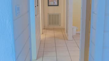A narrow hallway with white walls and a tiled floor, leading to a door with a vent.