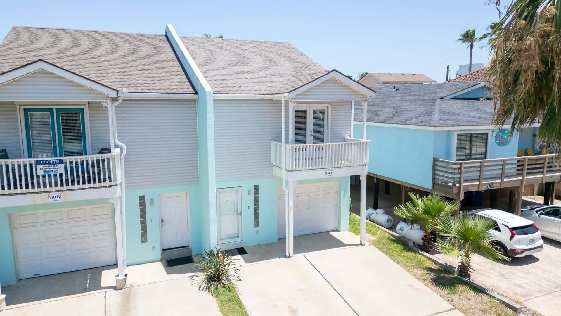 A two-story residential building with a garage, a balcony, and a small front yard with palm trees.