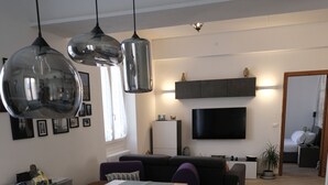 Una cuina moderna amb il·luminació penjant, una zona de menjador, un saló amb un sofà i un televisor fixat a la paret.