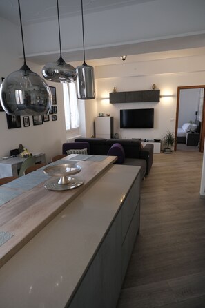 Eine moderne Küche mit Pendelleuchten, eine Essecke, ein Wohnzimmer mit Sofa und ein an der Wand befestigter Fernseher.