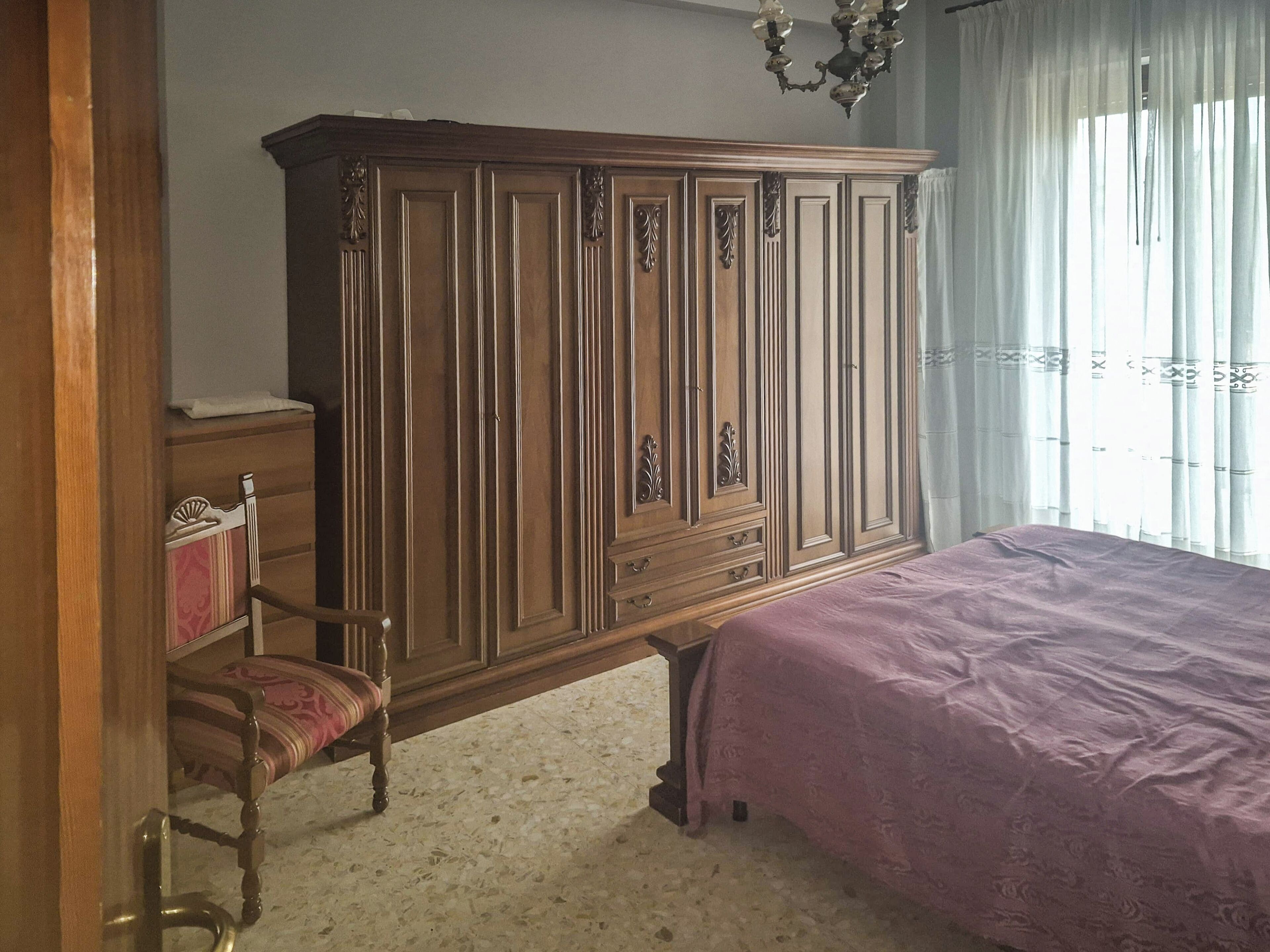 A bedroom with a wooden wardrobe, a bed with a purple bedspread, a chair, and a chandelier.