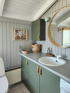 A bathroom with a white sink, green cabinets, a wooden-framed mirror, a wicker basket, and a framed floral painting on the wall. - Family holiday home on Als with sauna and shelter (Sydals)
