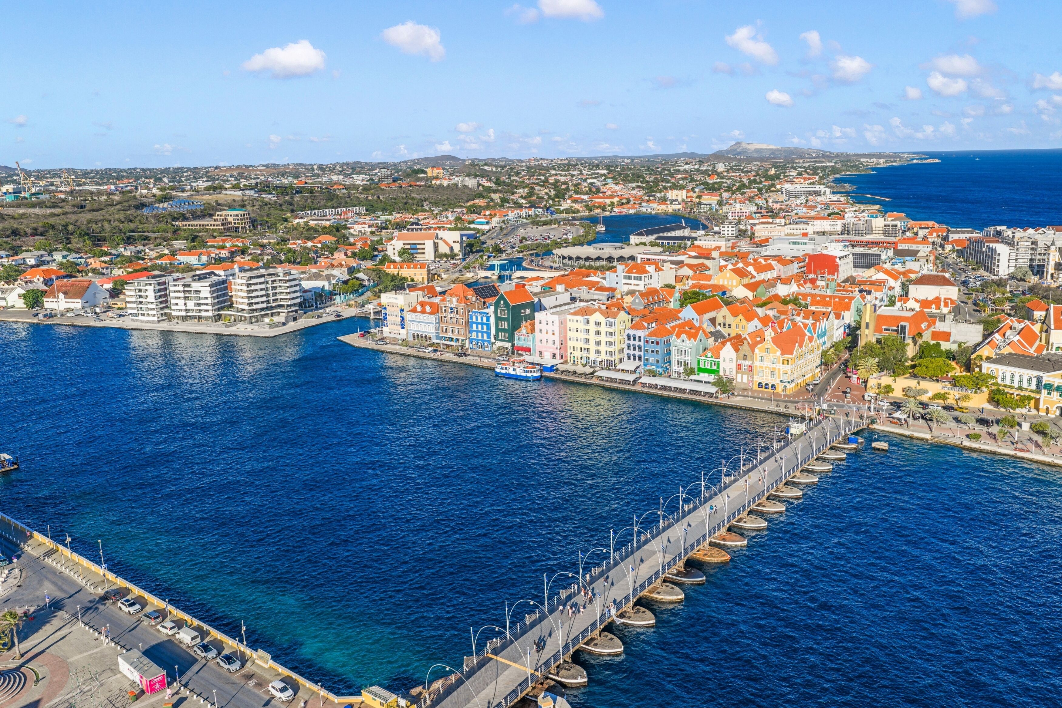Paesaggio costiero urbano con un molo prominente che si estende sull'acqua, circondato da edifici colorati e un cielo azzurro limpido.