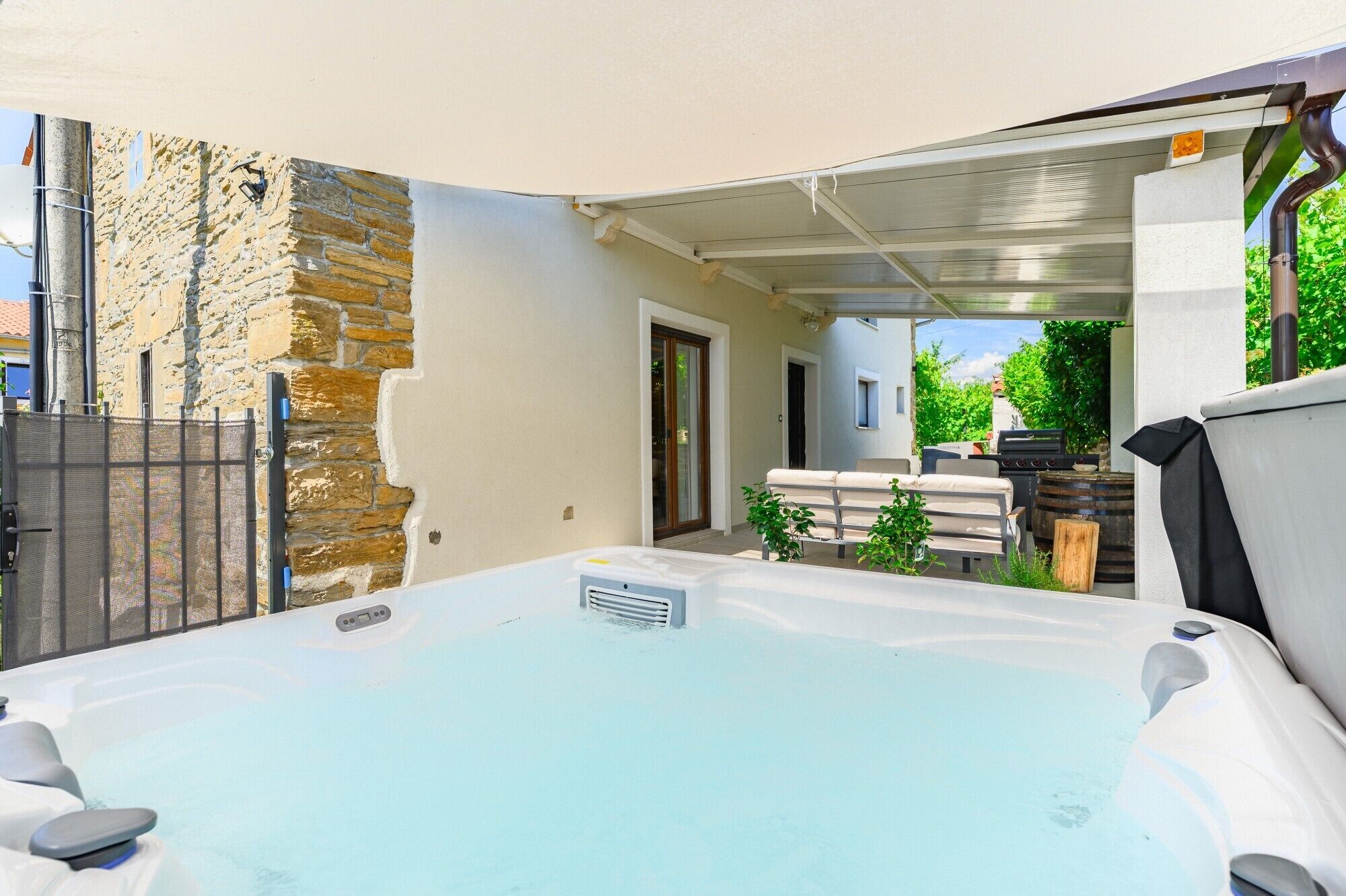 A hot tub in a covered outdoor area with a stone wall, a barbecue, and a patio with seating.