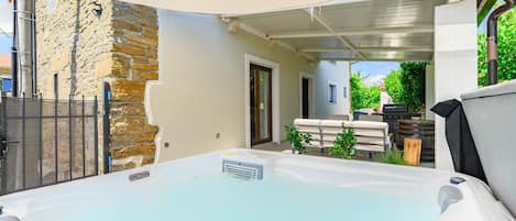 A hot tub in a covered outdoor area with a stone wall, a barbecue, and a patio with seating.