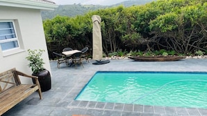 Una piscina con tumbonas de madera, una mesa y sillas, rodeada de vegetación y un muro de piedra.