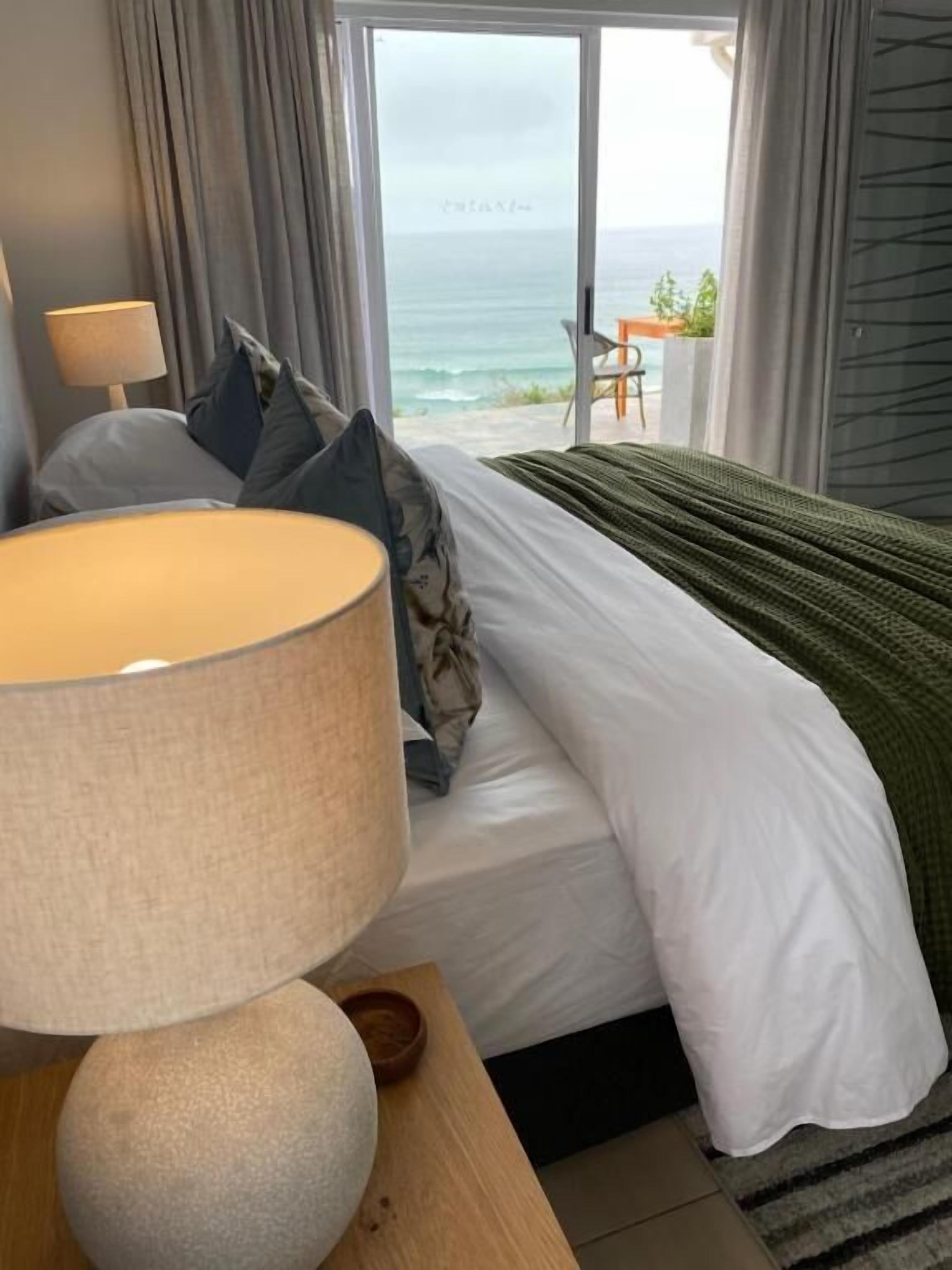 A bedroom with a bed, a lamp, a view of the ocean, and a chair by the window.