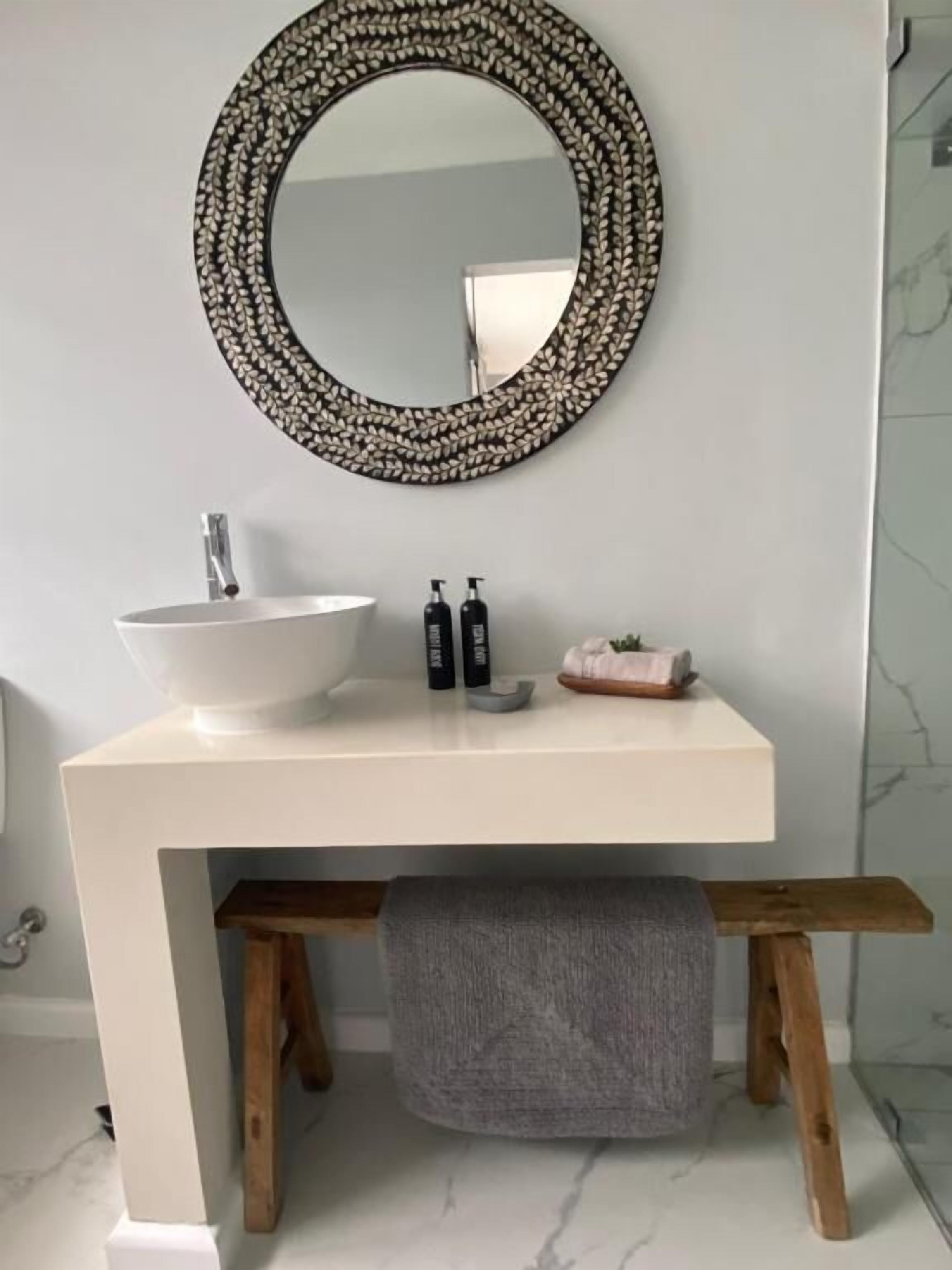 A bathroom with a round mirror, a white sink, a wooden bench, and a towel.
