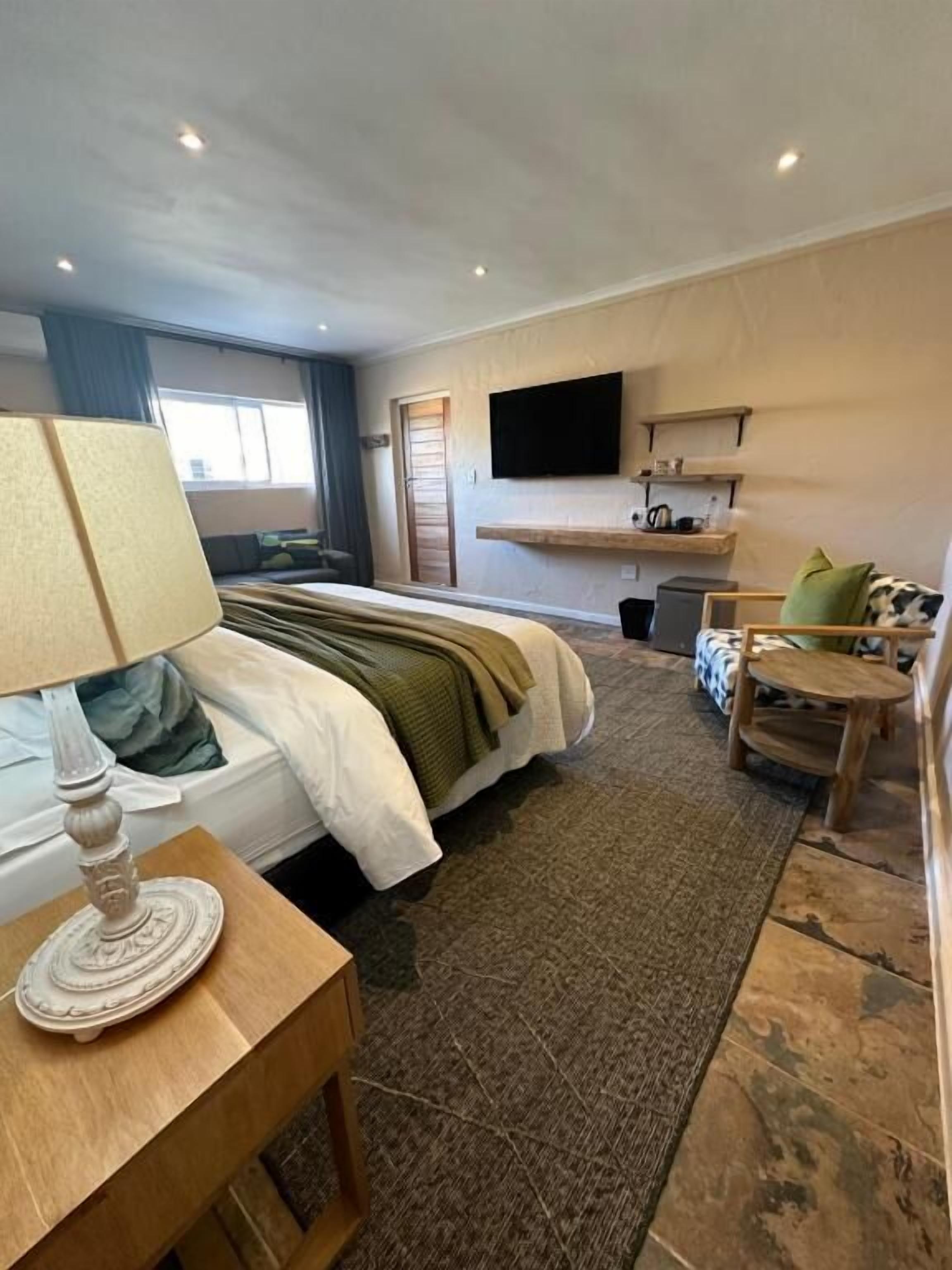 A hotel room with a bed, a bedside table with a lamp, a small round table, a TV mounted on the wall, and a window with curtains.