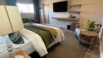 A hotel room with a bed, a bedside table with a lamp, a small round table, a TV mounted on the wall, and a window with curtains.