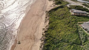 Vista aérea de una zona costera con playa, una carretera serpenteando a lo largo de la costa y un edificio con tejado inclinado.