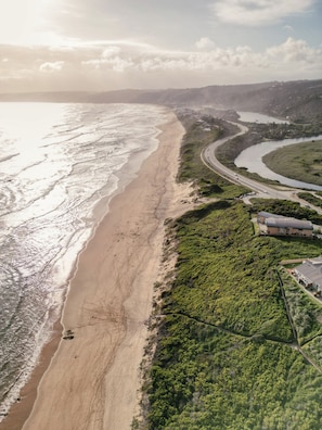 An aerial view of a coastal area with a beach, a road curving along the coast, and a building with a sloped roof. - Mes Amis Beach Guesthouse (Wilderness)