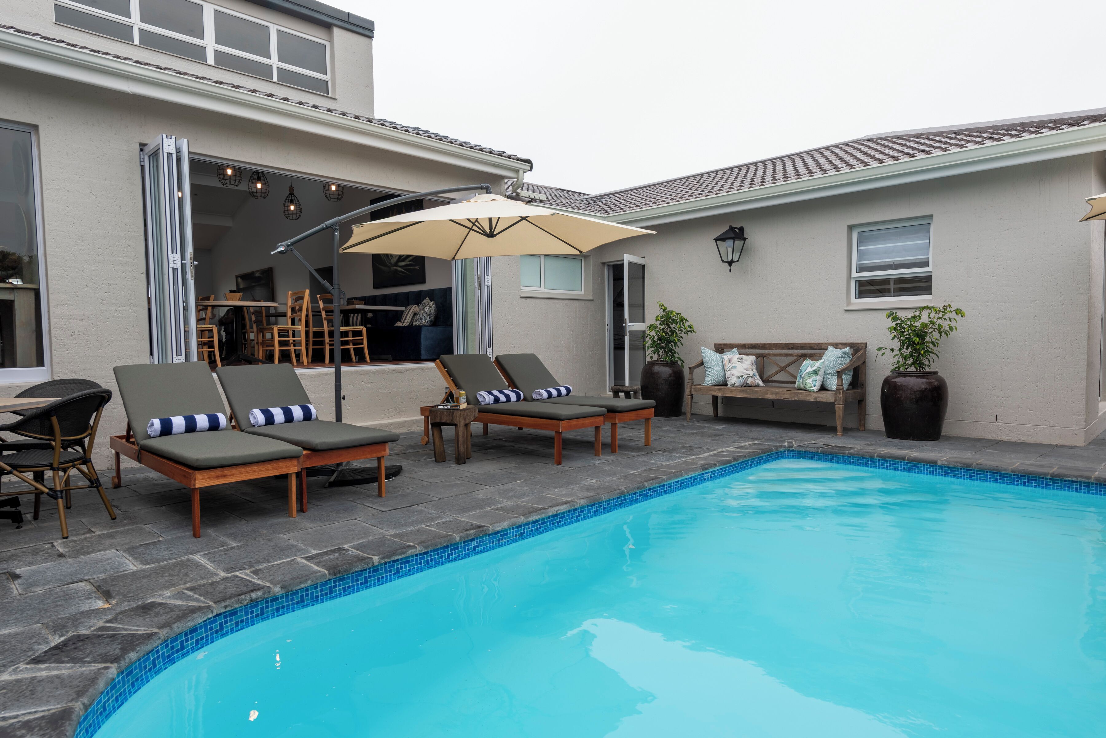 A swimming pool with lounge chairs and a table, a patio area with chairs, and a building with large windows and a door.