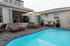 Uma piscina com espreguiçadeiras e uma mesa, uma área de estar com cadeiras, e um edifício com grandes janelas e uma porta.