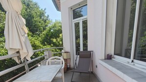 Ein Balkon mit Tisch und Stühlen, einer Liege und einem Fenster mit Blick ins Grüne.