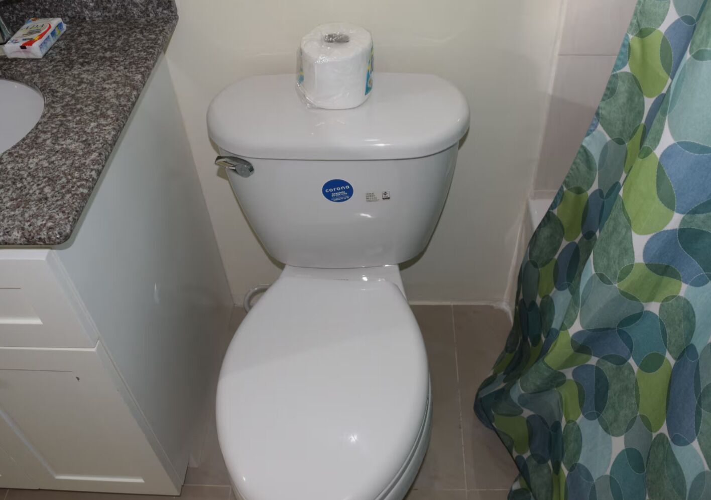 Un inodoro blanco con un adhesivo que dice 'corona', un rollo de papel higiénico y una encimera de baño con dispensador de toallas de papel.