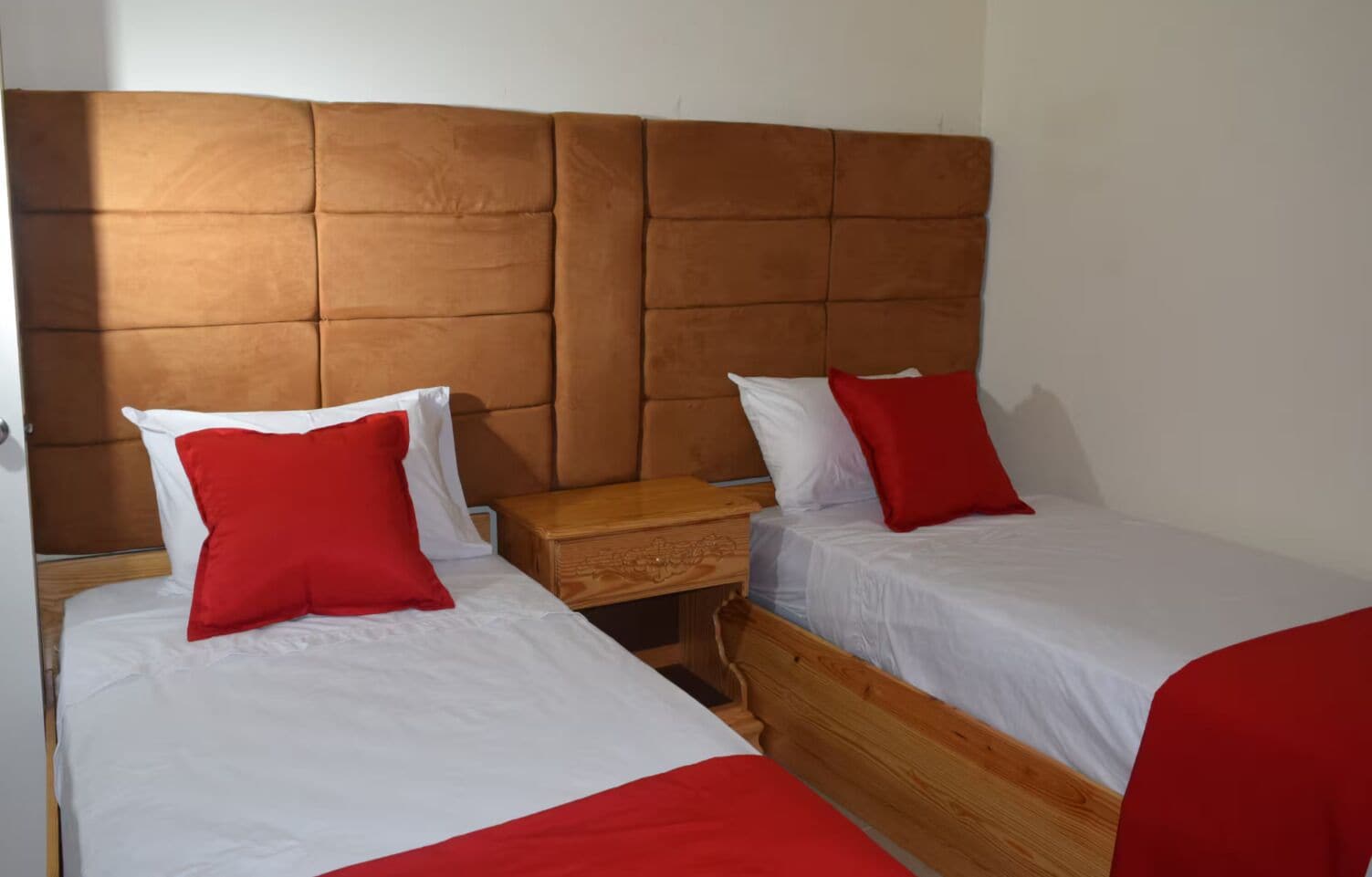 A room with two beds, each with white bedding and red pillows, a wooden nightstand, and a brown padded headboard.
