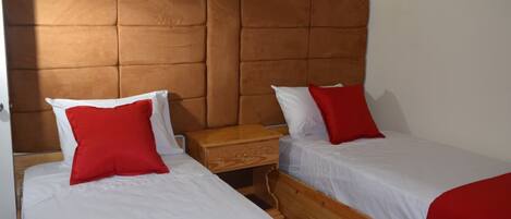 A room with two beds, each with white bedding and red pillows, a wooden nightstand, and a brown padded headboard.