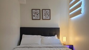 A neatly made bed with white linens, a dark headboard, and two framed botanical prints on the wall.