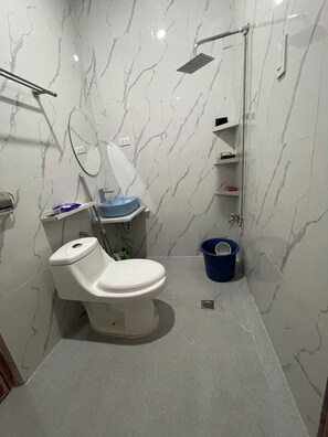 A bathroom with a toilet, a shower, a mirror, and a blue bucket. - LEVA Suites- At Bagontaas, close to Bukidnon’s Mountains, Lakes & Nature Escapes (Valencia City)