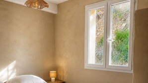 Un dormitorio con cama, lámpara de noche y ventana con vista a la vegetación.