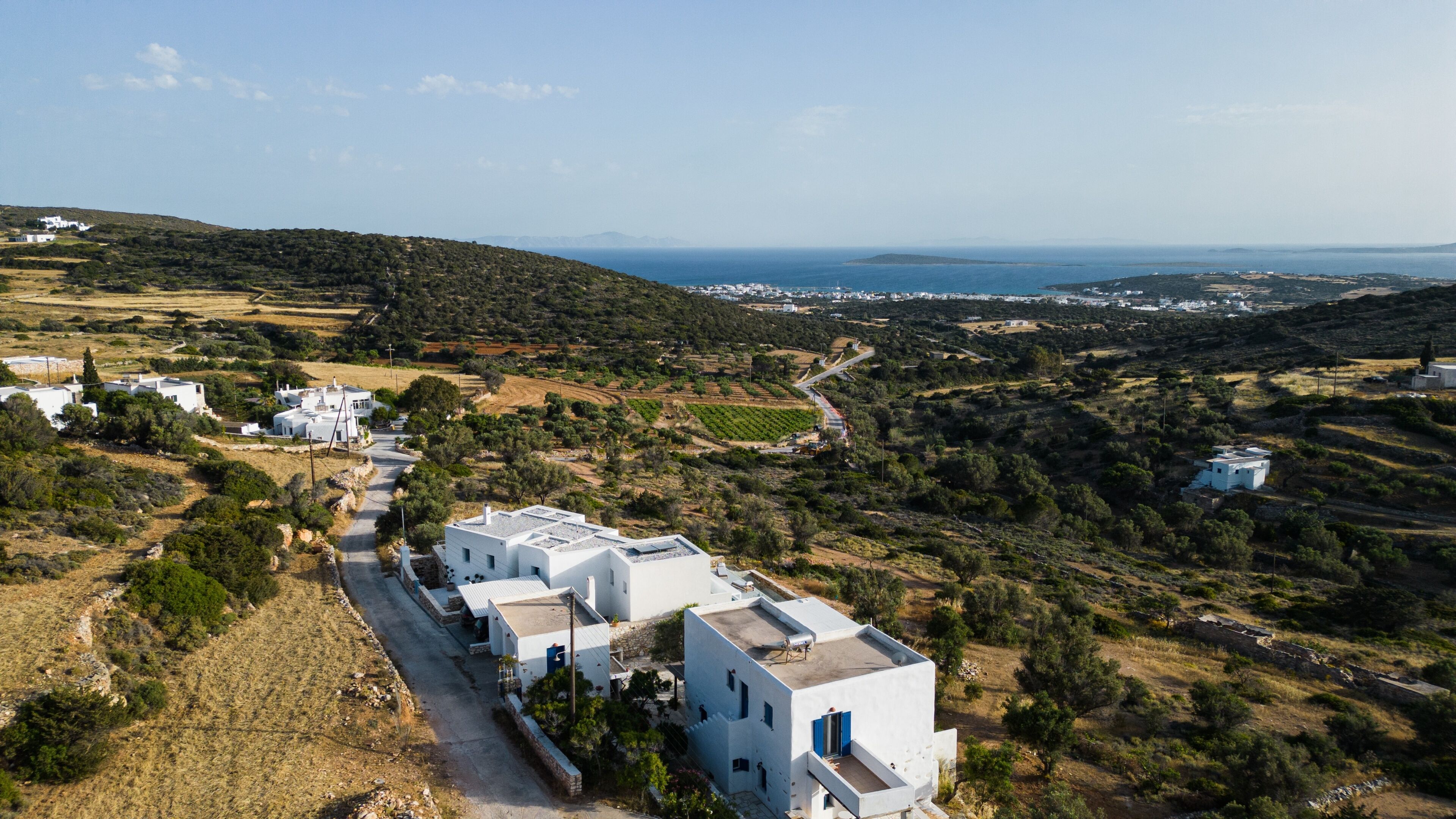 A coastal village with white buildings, a road, and a view of the sea and distant mountains.