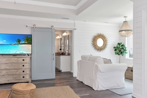 A modern living room with a flat-screen TV, a white sofa, a dining area with a kitchen, and a large window with a view. - 🌊 Arlanymor | Windmark Beach | Private Pool, Volleyball + Beach Access (Windmark)