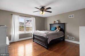 Un dormitorio con cama, ventilador de techo, ventana con cortinas y vista a una casa.