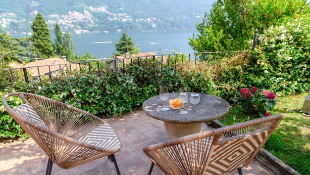 Eine Terrasse mit Korbstühlen und einem Steintisch, mit Blick auf einen See und Hügel.
