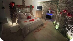 Un dormitorio con pared de piedra, una cama con pétalos de rosa y un televisor sobre un mueble de madera.