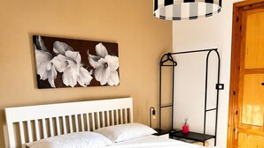 Ein ordentlich bezogenes Bett mit weißen Laken, ein Nachttisch mit Lampe und ein Wandbild mit weißen Blumen.