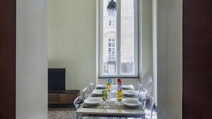 Ein Essbereich mit Tisch für vier Personen, einem Fernseher im Hintergrund und einem Fenster mit Blick auf Gebäude.