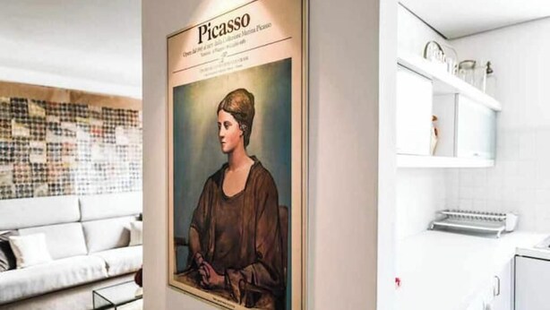 Ett inramat konstverk på en vägg, en vit soffa och en köksdel med en tvättmaskin.