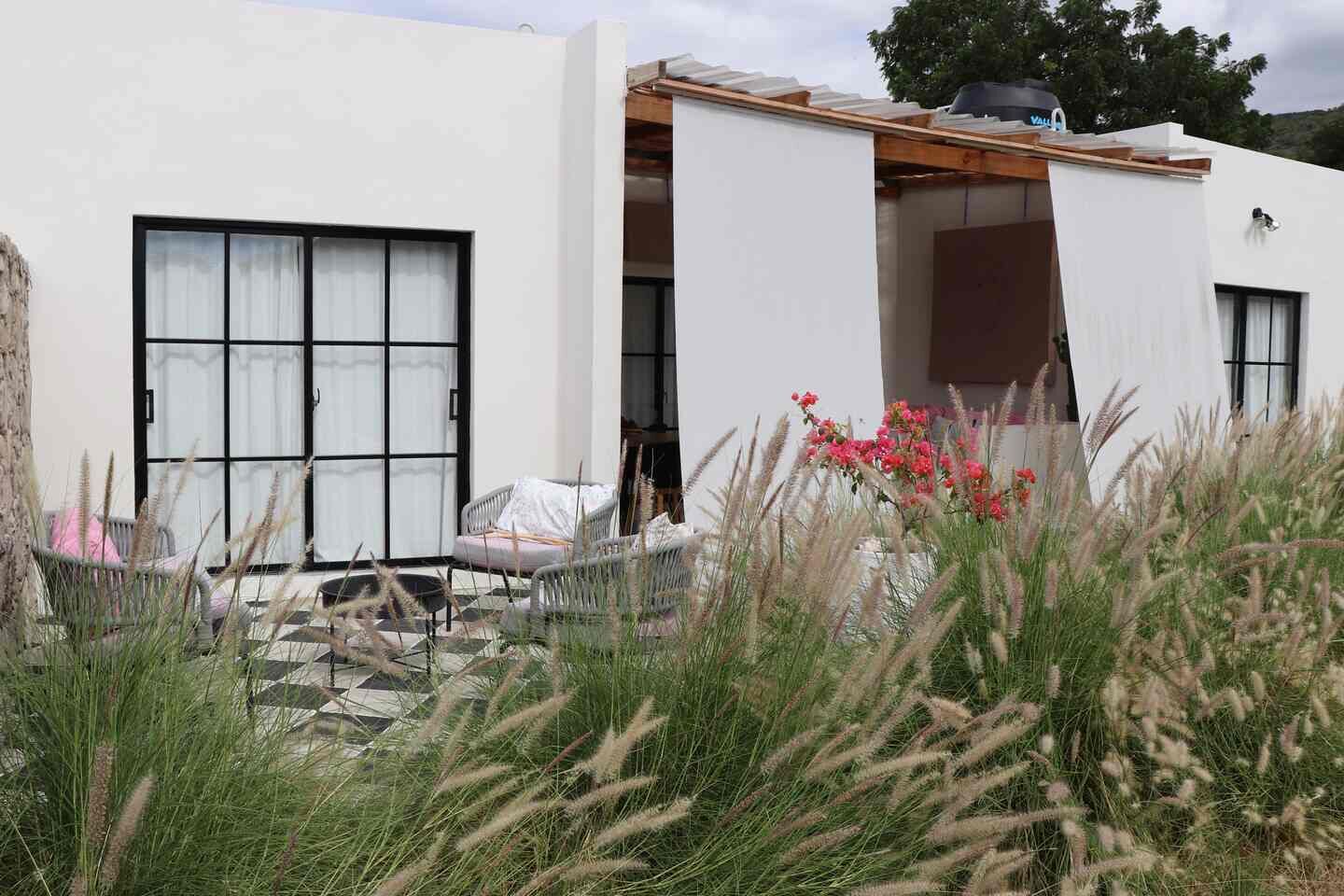 Een modern huis met een groot raam, een overdekt terras en een tuin met hoge grassen en roze bloemen.