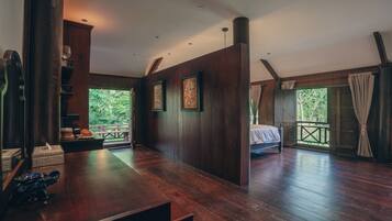 A room with a wooden floor, a bed, a bench, and a view of greenery outside.